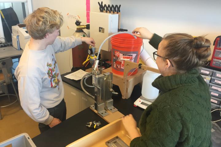 A photo of two students with a red bucket in between them and hoses going in and out of the bucket