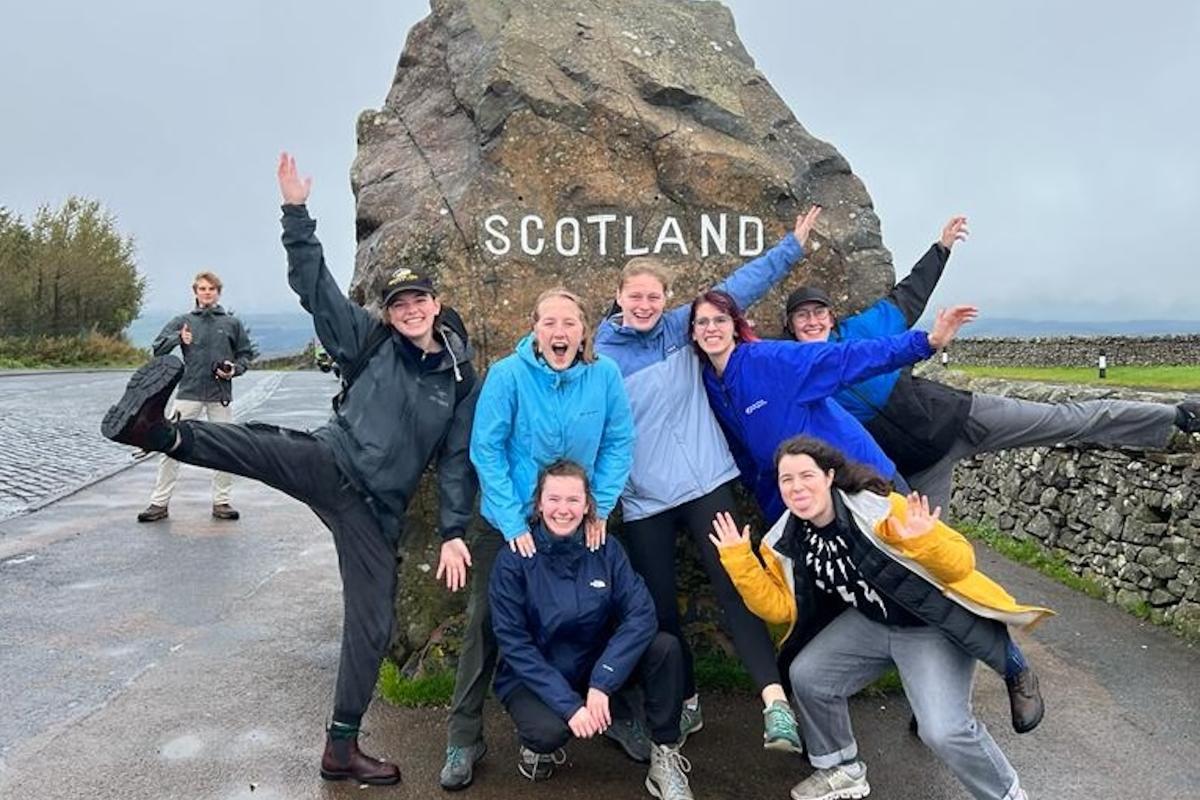 A group of college students pose for a joyful photo in front of a large rock with the words "SCOTLAND' across it in white lettering. A group of college students pose for a joyful photo in front of a large rock with the words "SCOTLAND' across it in white lettering.